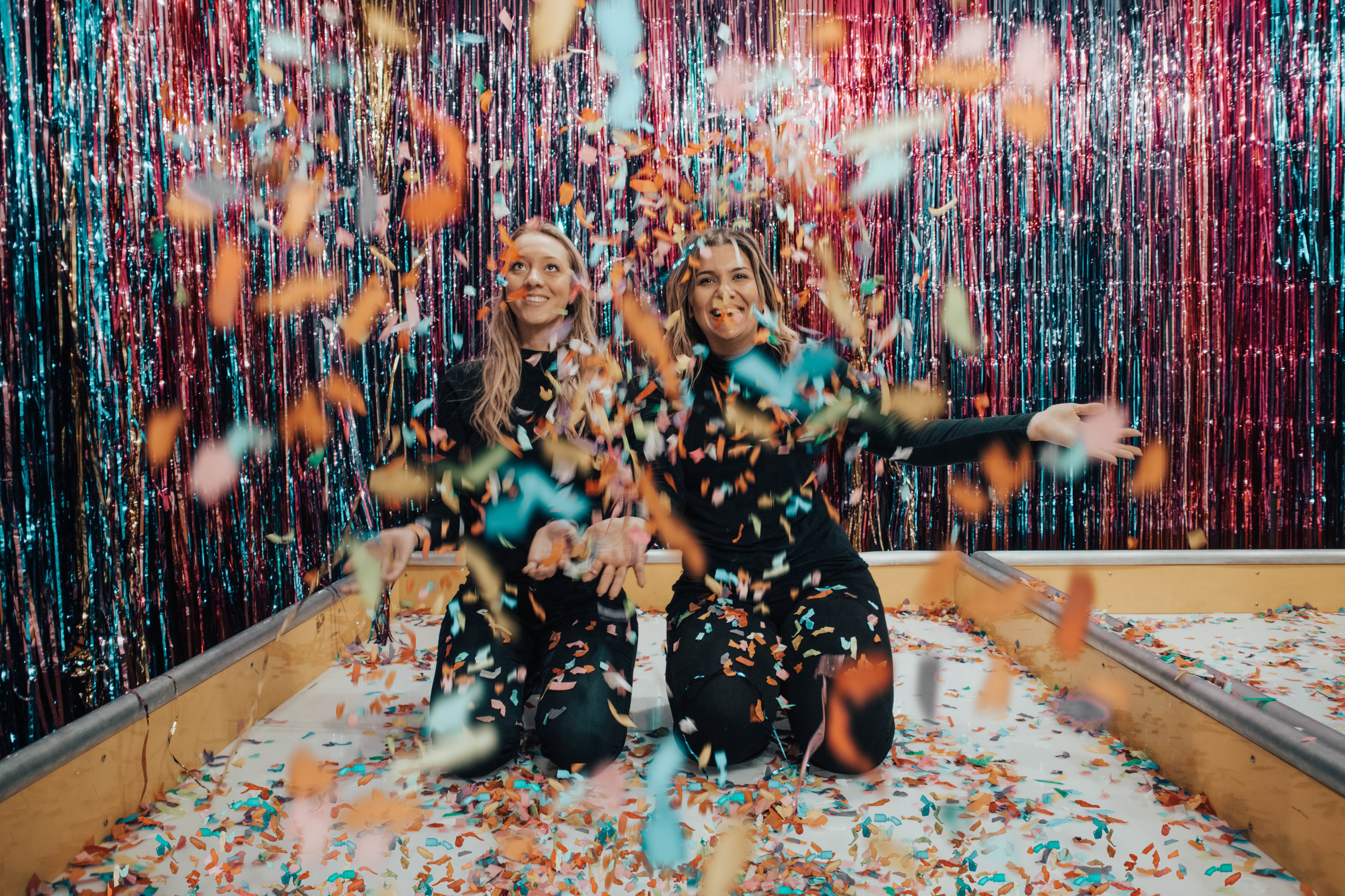 Two woman on their knees  smiling and throwing confetti 